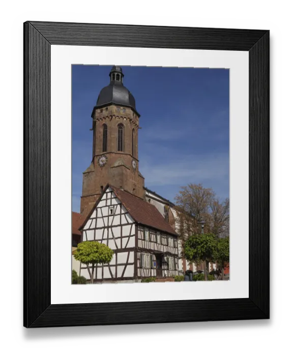 Framed Print : A church tower towers over a traditional half-timbered house under a bright blue sky