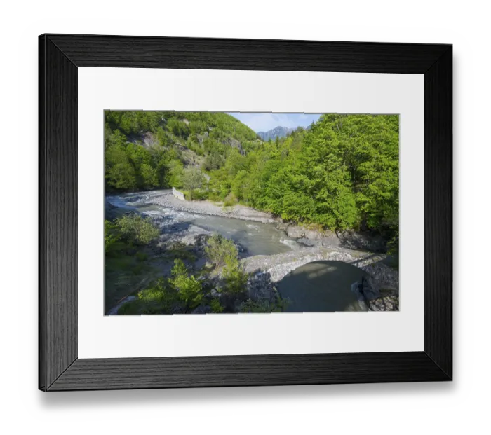 Framed Print : A clear river flows under an old stone bridge through a green