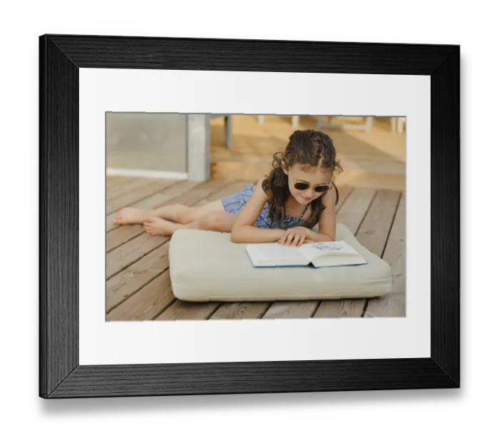 Framed Print : Girl with sunglasses sits on cushion on wooden terrace and reads a book