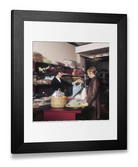 Framed Print : Shopping in the 1960s. Interior of a fashion shop where a woman is showing a female customer a swimming costume