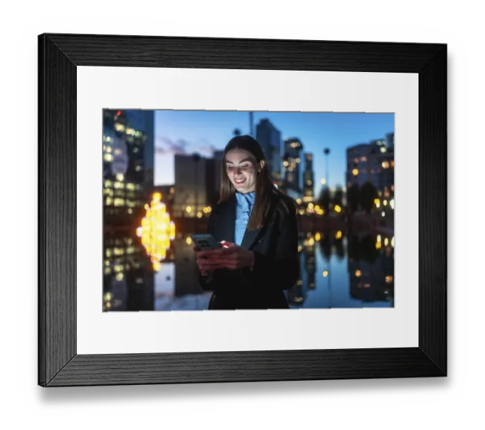 Framed Print : Young professional woman smiling happily while interacting with her mobile phone against a blurred city background with