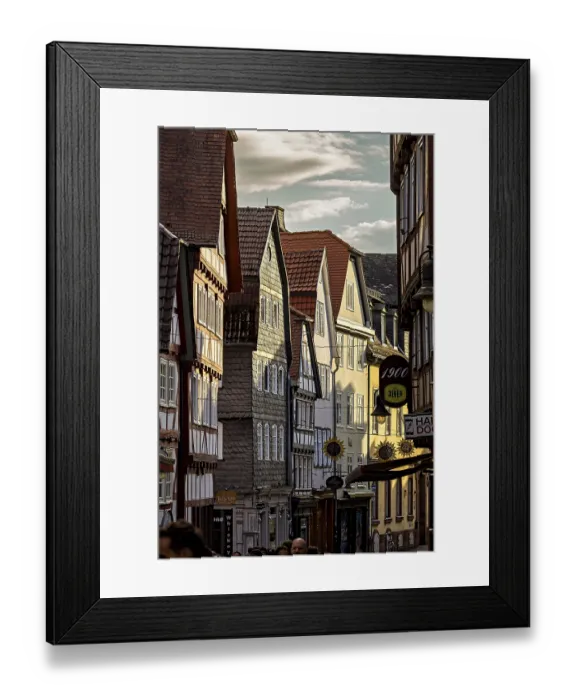 Framed Print : Narrowly built street with historic half-timbered houses and people passing by in the evening light