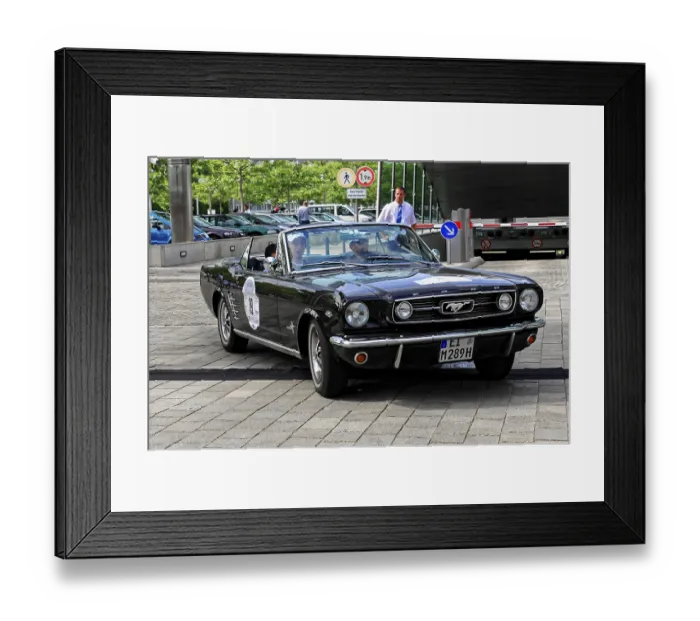Framed Print : Black vintage convertible with driver and passenger on paved road in front of modern buildings