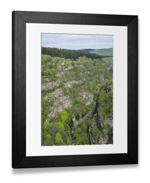 Framed Print : A green gorge with rocks and trees crossed by a small river under a cloudy sky