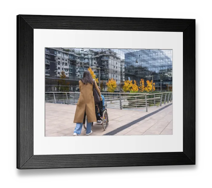Framed Print : Woman in a brown coat pushing a young person in a wheelchair along a modern urban sidewalk