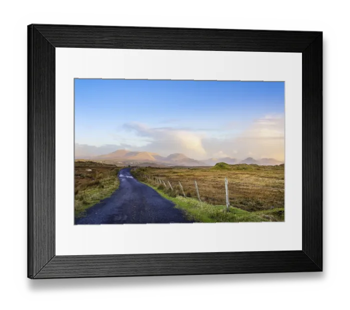 Framed Print : Empty country road between meadows leading to mountains in far distance