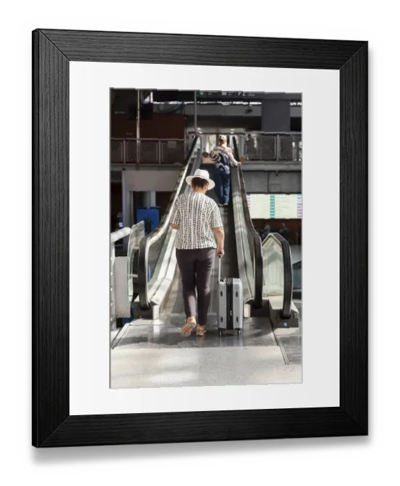 Framed Print : Senior woman in white hat pulling suitcase up an escalator inside modern transport terminal
