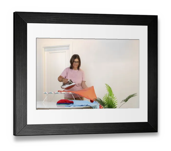 Framed Print : Woman standing smiling while ironing a colorful t shirt on an ironing board