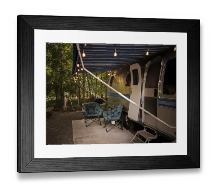 Framed Print : An Airstream camper trailer with string lights and chairs set up in a forest