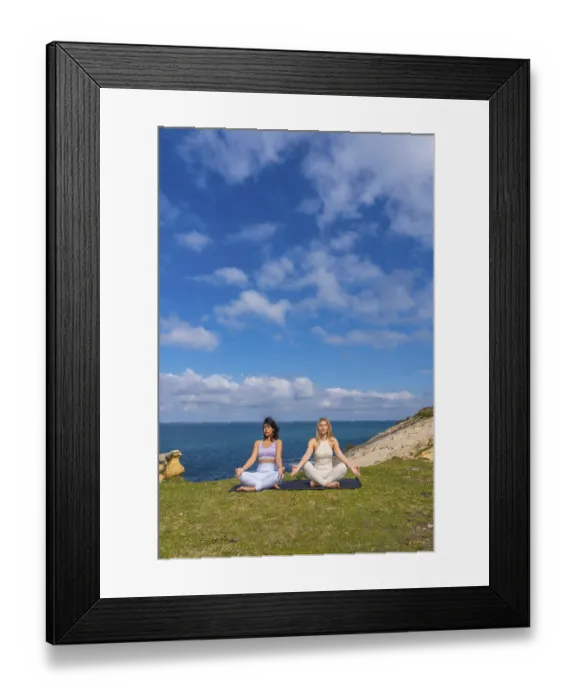 Framed Print : Two women practicing meditation and yoga in a serene lotus pose on a yoga mat overlooking the ocean from a grassy cliff
