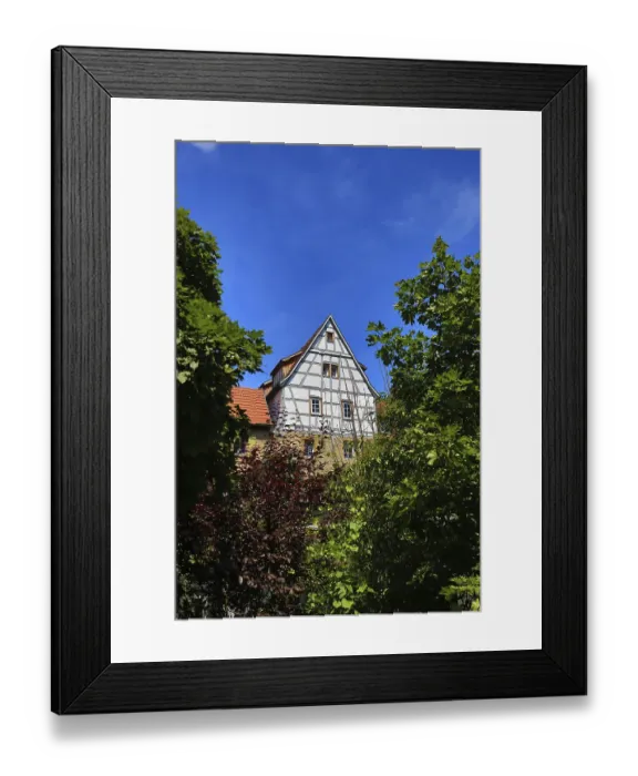 Framed Print : Half-timbered house towering between green trees in the blue sky Framed Print : Half-timbered house towering between green trees in the blue sky