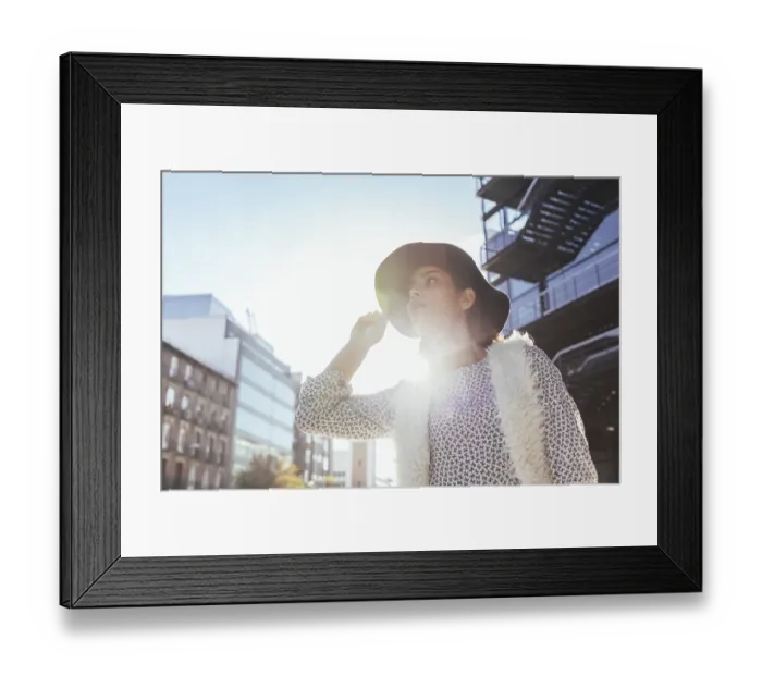 Framed Print : A fashionable woman in a wide-brimmed hat and patterned blouse stands illuminated by sunlight in an urban environment