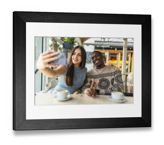 Framed Print : Smiling diverse couple making a peace sign gesture while taking a selfie with a smartphone at a cozy coffee shop