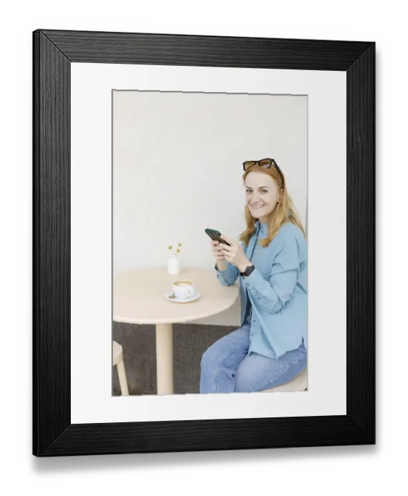 Framed Print : A smiling woman enjoys coffee while using her smartphone at a cafe in Warsaw She wears casual clothing and sunglasses
