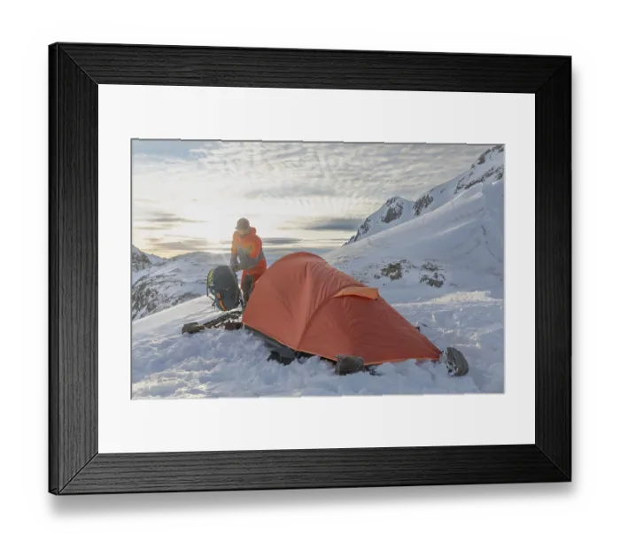 Framed Print : A ski tourer sets up near a bright orange tent amidst snowy mountains at sunrise