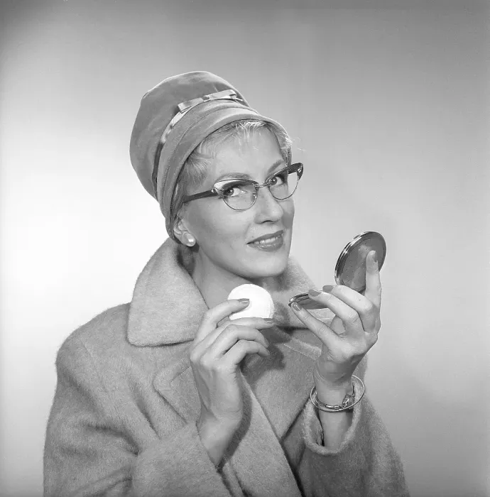 Framed Print : 1950s makeup. A woman is looking at herself in her pocket mirror and improves on her makeup. She is wearing a fashionable hat, typical 1950s glasses and a coat. 1958 Kristoffersson ref CB96-5