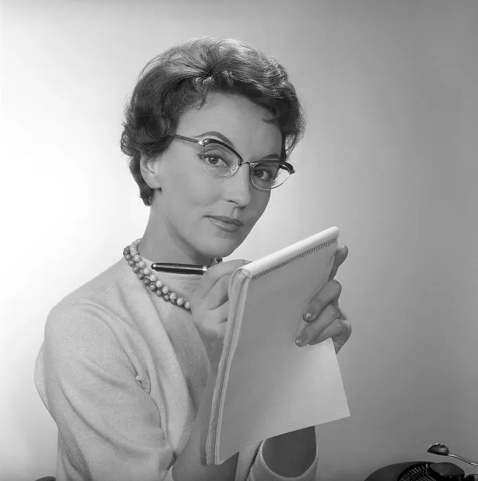Framed Print : In the 1950s. A woman at a desk with a typewriter visible holds a notepad and writes in it with a pen