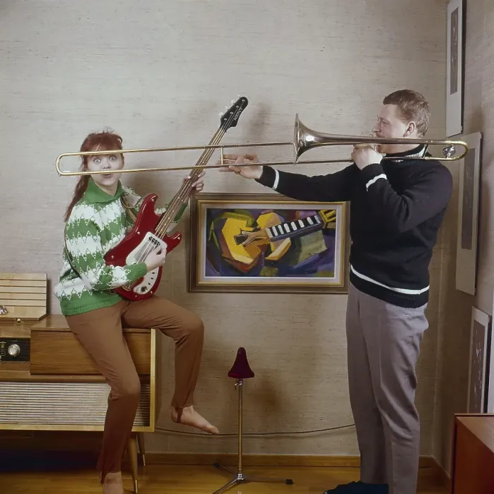 Framed Print : In the 1960s. A couple is seen playing the trombone and an electric guitar, looking happy and playful. To the left a radio gramophone. A combined music furniture that could play records and radio in the same unit. Often a large piece of furniture Framed Print : In the 1960s. A couple is seen playing the trombone and an electric guitar, looking happy and playful. To the left a radio gramophone. A combined music furniture that could play records and radio in the same unit. Often a large piece of furniture