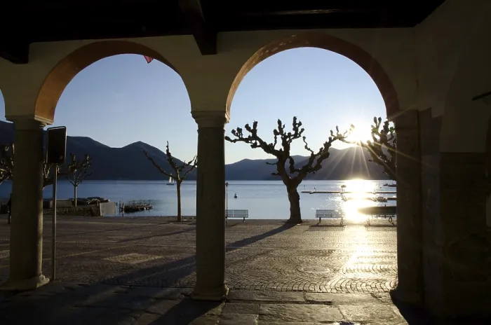 Framed Print : Arch and Bare Tree on the Waterfront to Lake Maggiore with Mountain and Sunbeam in Sunset in Ascona