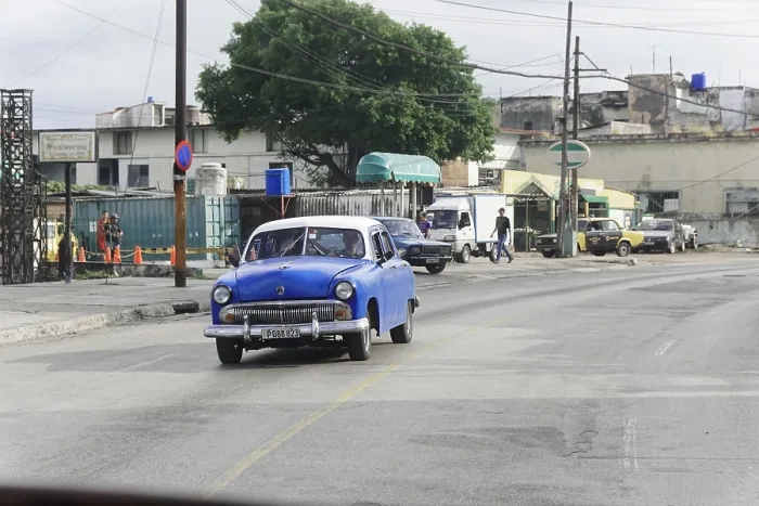 Framed Print : A blue classic car on a road surrounded by buildings and trees
