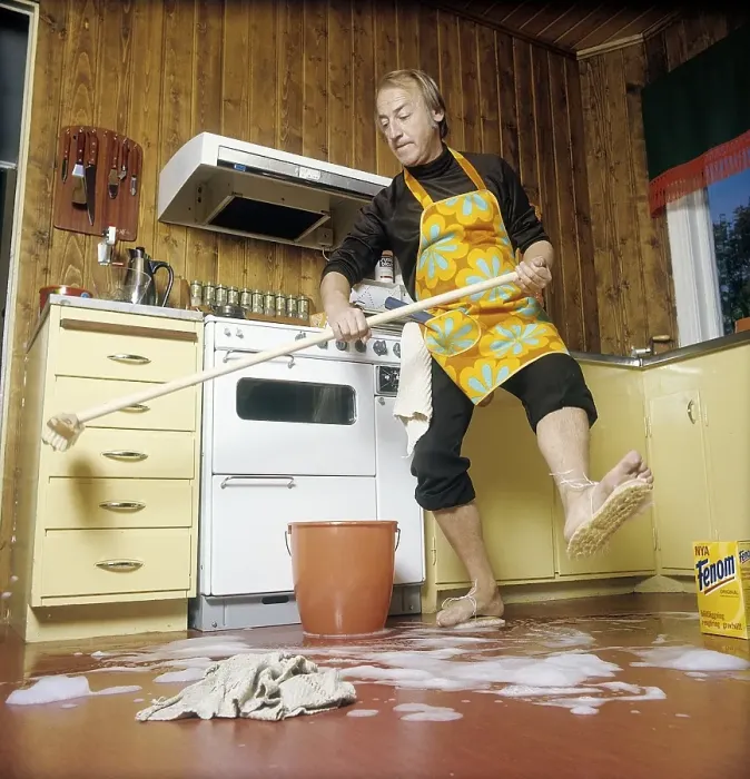 Framed Print : Cleaning day in the 1970s. An interior of a kitchen where a man is seen cleaning the floor