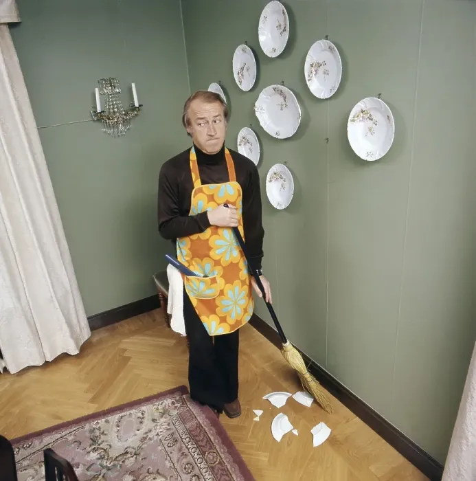 Framed Print : Cleaning day in the 1970s. An interior of a man cleaning the floor. Bits of a plate that used to hang on the wall is seen on the floor, he must have torn it down with the handle