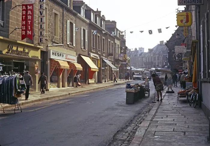 Framed Print : Eine Reihe von Geschaeften mit Menschen, die die zum Marktplatz fuehrende Strasse buersten, vermutlich in Quimper, Bretagne, Frankreich, 1974 Framed Print : Eine Reihe von Geschaeften mit Menschen, die die zum Marktplatz fuehrende Strasse buersten, vermutlich in Quimper, Bretagne, Frankreich, 1974