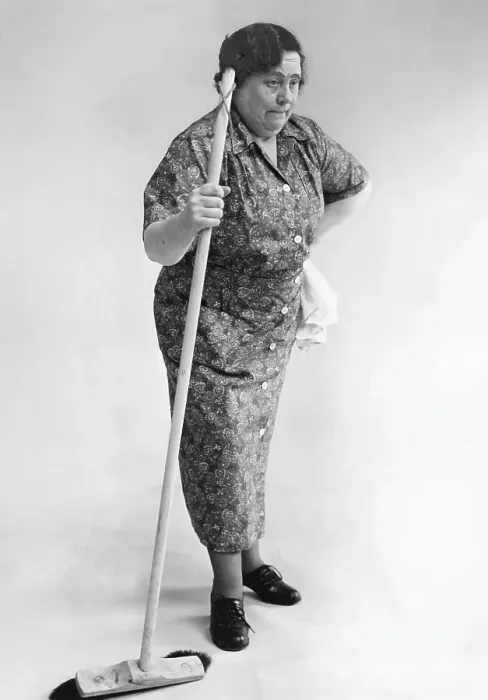 Framed Print : Elderly cleaning lady looking worn and slightly forward as if she has a pain in her back. She does not look very happy standing there leaning against a brush. 1940s
