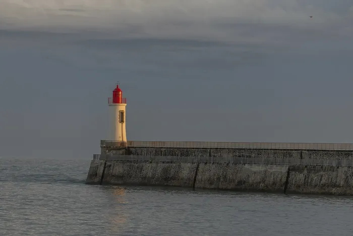 Framed Print : A lighthouse stands on a pier and casts its light over calm waters at dusk