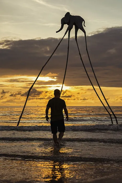 Framed Print : A man stands in the surf facing a surreal elephant sculpture with long legs