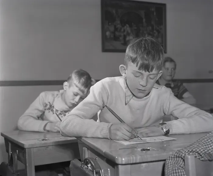 Framed Print : School in the 1940s. The interior of a classroom with a class of children sitting neatly at their desks. A boy is sitting at the front of the class, writing with an ink pen. The school desks had a lid that opened