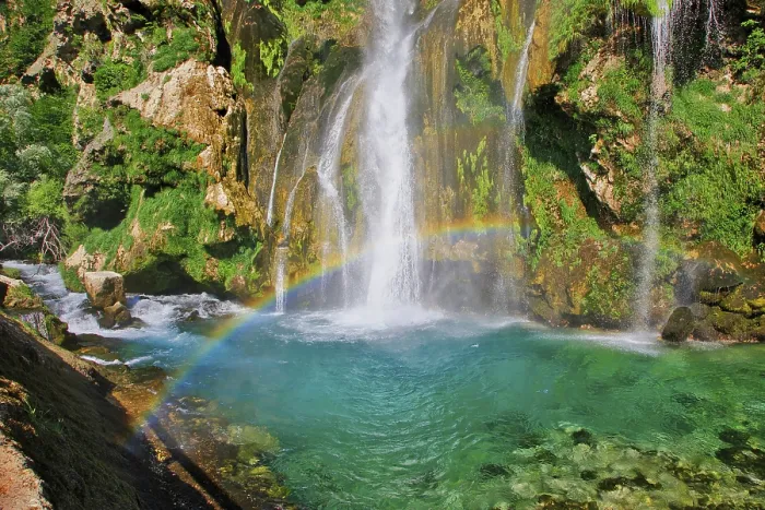 Framed Print : Waterfall with rainbow, surrounded by rocks and green foliage under a clear sky
