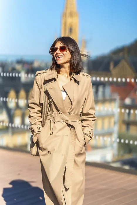 Framed Print : A woman wearing a tan trench coat is smiling and walking on a bridge