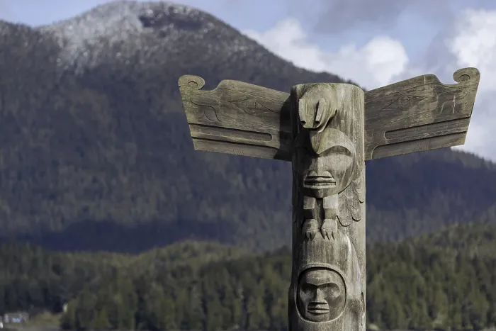 Framed Print : Wooden totem pole stands in front of scenic mountain and forest backdrop under a cloudy sky in Tofino