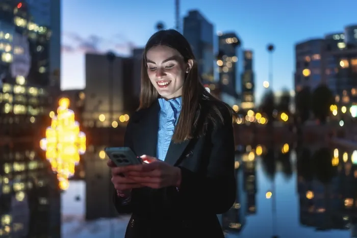 Framed Print : Young professional woman smiling happily while interacting with her mobile phone against a blurred city background with