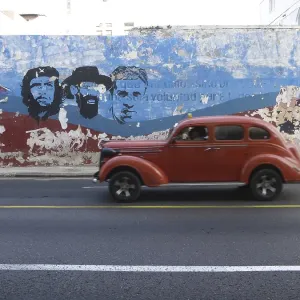 American vintage car driving along a wall with paintings of Fidel Castro and Che Guevara