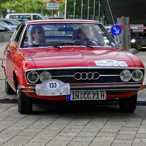 AUDI, A red vintage car drives on cobblestones with two people in the car