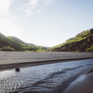 Beach with a flowing river, surrounded by hills and mountains under a blue sky Beach with a flowing river, surrounded by hills and mountains under a blue sky