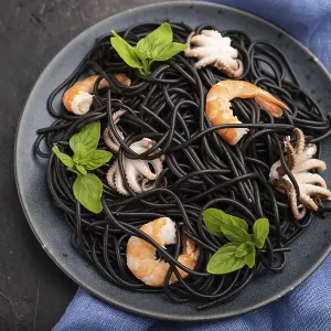 Black cuttlefish ink pasta with shrimps or prawns and small octopuses on black concrete background and blue textile Black cuttlefish ink pasta with shrimps or prawns and small octopuses on black concrete background and blue textile