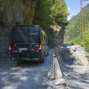 A black van drives on a narrow mountain road surrounded by rocks and thick vegetation A black van drives on a narrow mountain road surrounded by rocks and thick vegetation