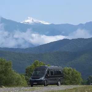 A black van on a road in a mountain valley with snowy peaks and clouds