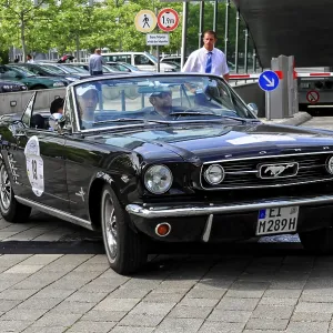 Black vintage convertible with driver and passenger on paved road in front of modern buildings