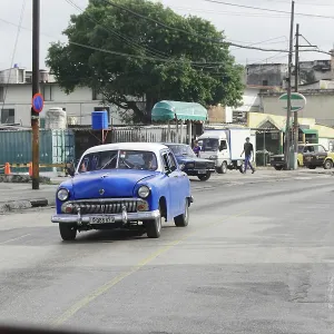 A blue classic car on a road surrounded by buildings and trees