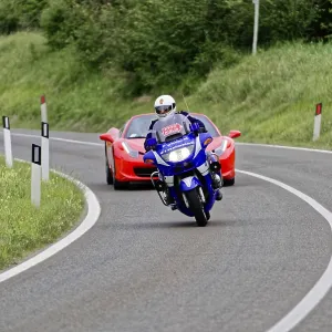 Blue motorbike takes a bend in front of a red Ferrari convertible on a country road Blue motorbike takes a bend in front of a red Ferrari convertible on a country road