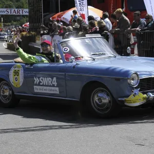 A blue open-top vintage rally car with a smiling driver in front of spectators A blue open-top vintage rally car with a smiling driver in front of spectators