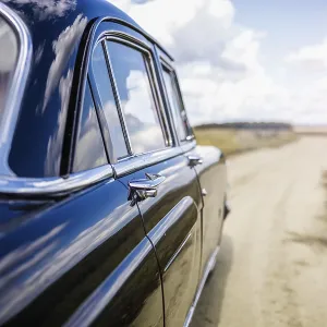 A blue vintage car on a rural road under a cloudy sky