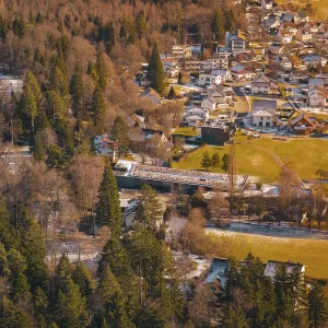 A bridge over a river surrounded by trees leads to a village in a snowy landscape