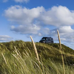A campervan stands on a grassy dune under a blue sky with clouds A campervan stands on a grassy dune under a blue sky with clouds