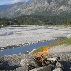 A campfire in a rocky landscape with mountains and a riverbed in the background