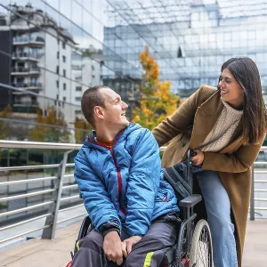 Caregiver woman pushes smiling young man in wheelchair along a city sidewalk